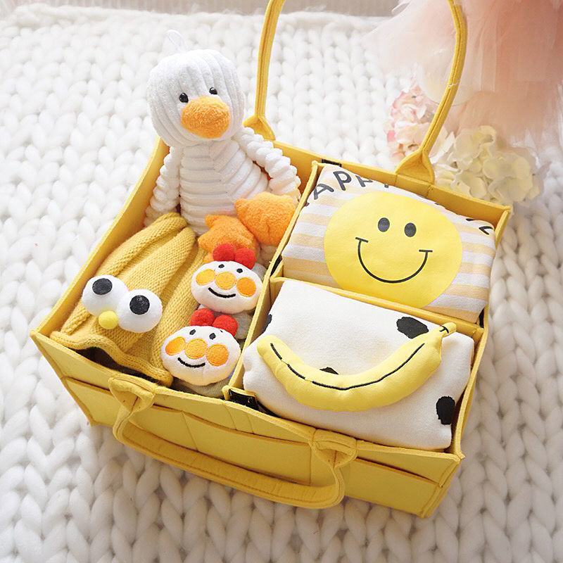 Set of baby items including a plush toy, bib, and socks in a yellow basket on a white textured background.